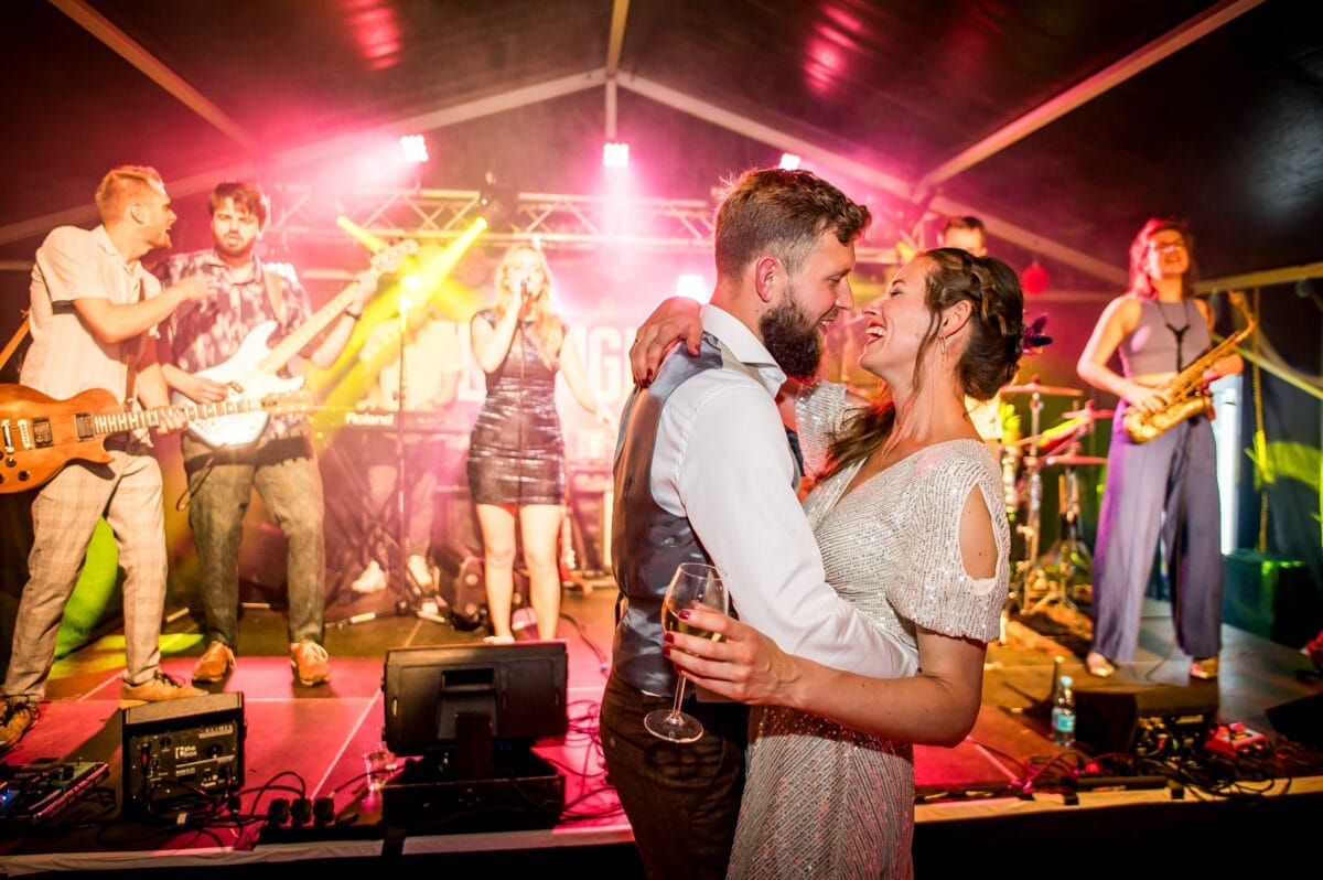 Stel een dansende stel voor een live band op een kleurrijke verlicht podium met feestelijke sfeer, wijnglas in de hand, genoten van levendige muziek en vrolijke ambiance tijdens het evenement.