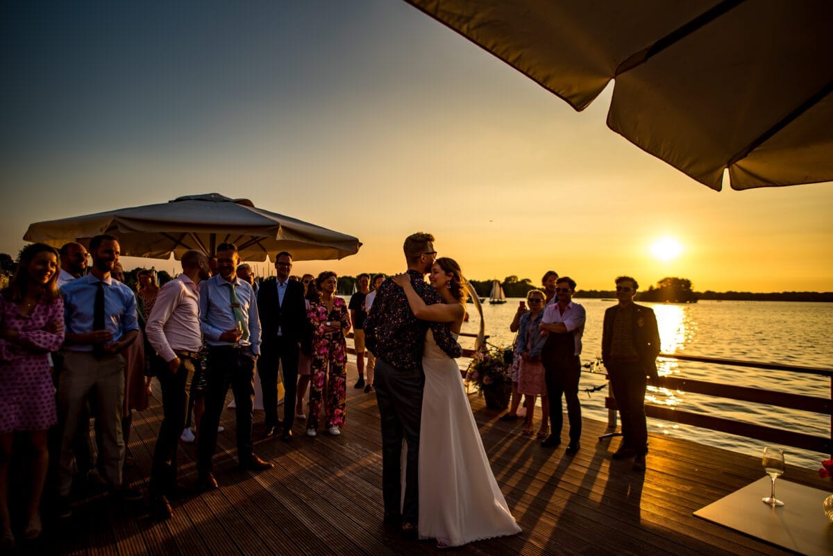 Stelletjesdans op houten steiger bij zonsondergang, omringd door lachende gasten, grote parasols, bloemendecoratie en glazen aan het serene meer in warme gouden avondzon.