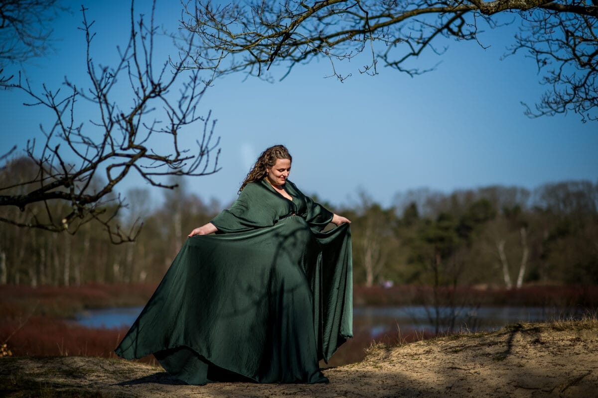 Zwangerschapsfotoshoot op zandgrond bij meer, vrouw in lange donkergroene jurk poseert tussen kale takken en bos, stralende glimlach onder betrouwbare blauwe lucht.