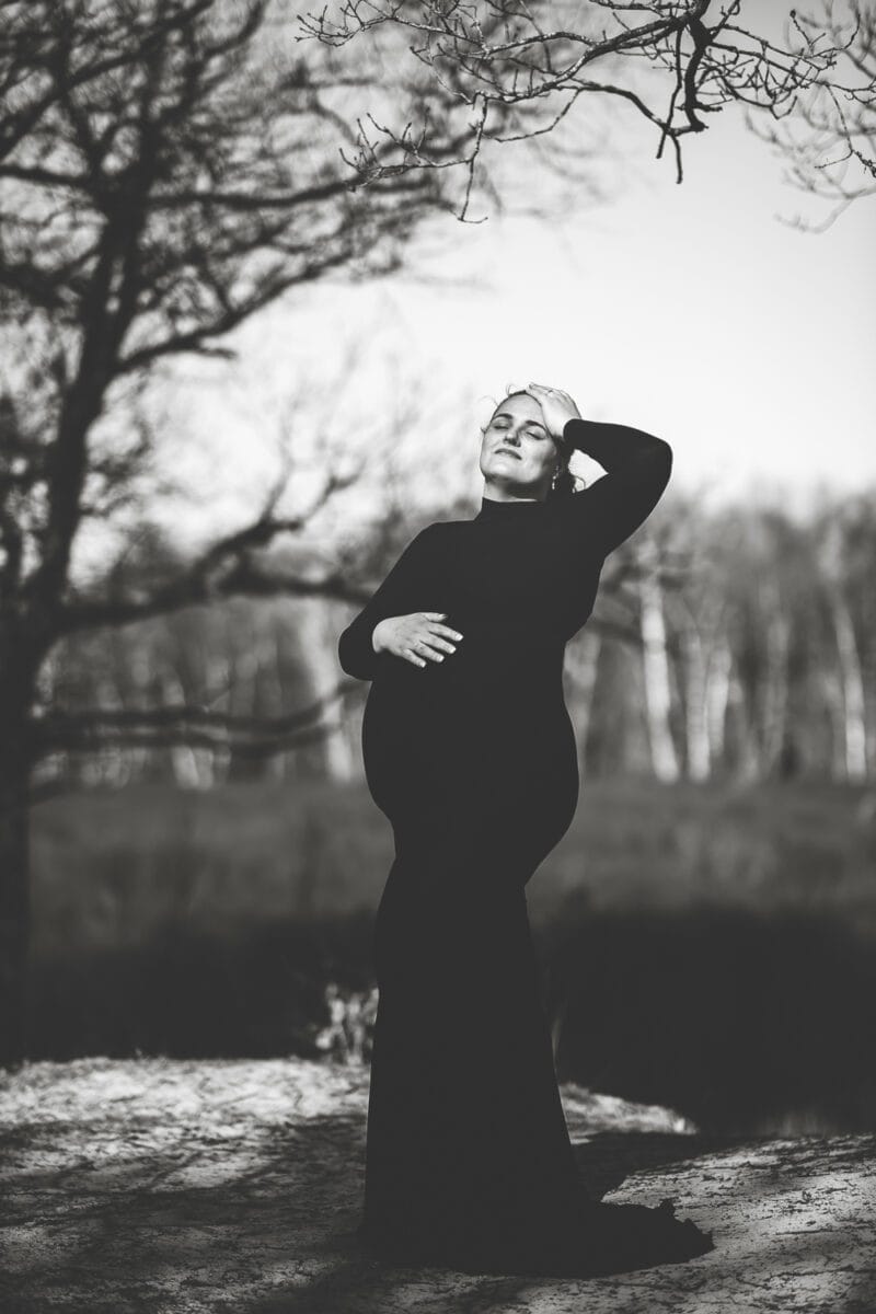 Zwangere vrouw in lange zwarte jurk poseert buiten voor een zwangerschapsfotoshoot; hand op buik, hand op hoofd, bomen en onscherpe achtergrond; zwart-wit zwangerschapsfotografie.