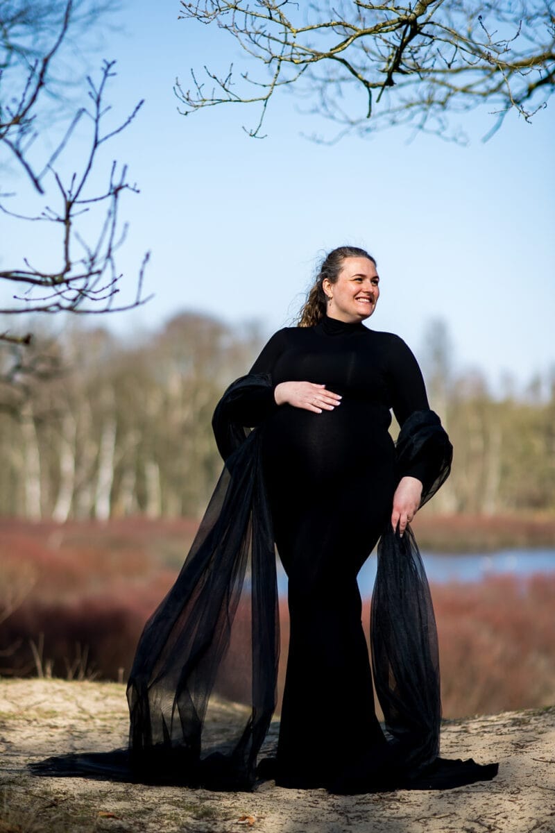 Zwangerschapsfotografie buiten: zwangere vrouw in elegante zwarte jurk omarmt haar buik tussen boerenkool boomtakken, zachte achtergrond met bomen en lucht.