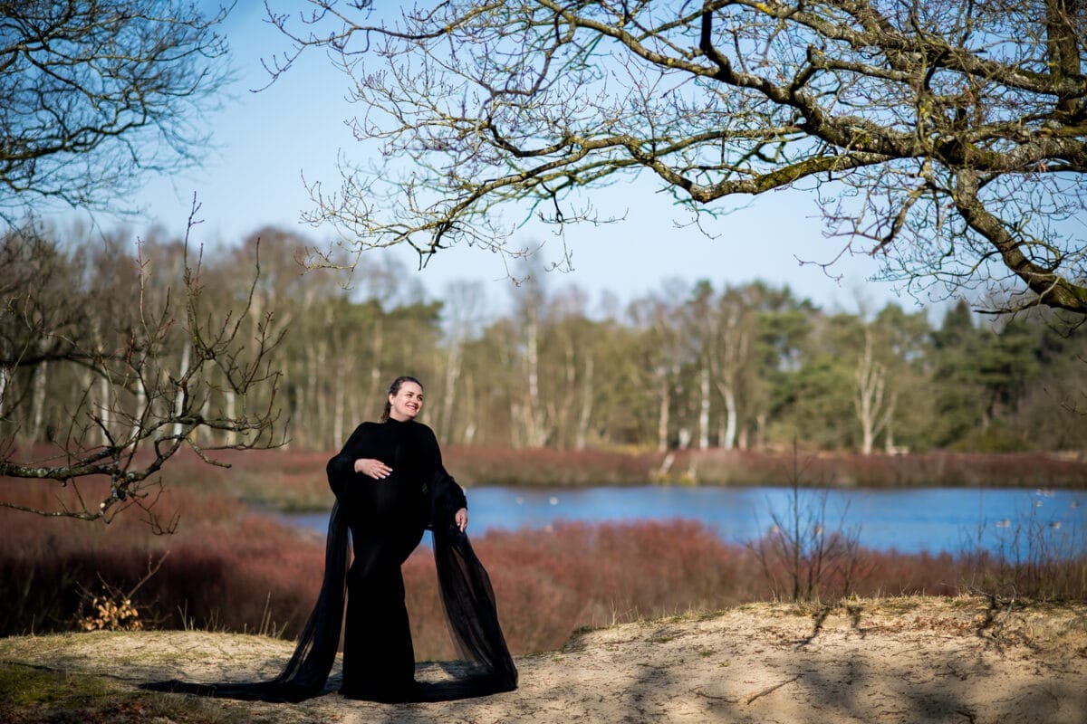 Vrouw in lange zwarte jurk poseert lachend op zandgrond bij een meer, omringd door kale bomen en herfststruiken, tijdens een zwangerschapsfotoshoot onder een betrouwbare blauwe lucht.