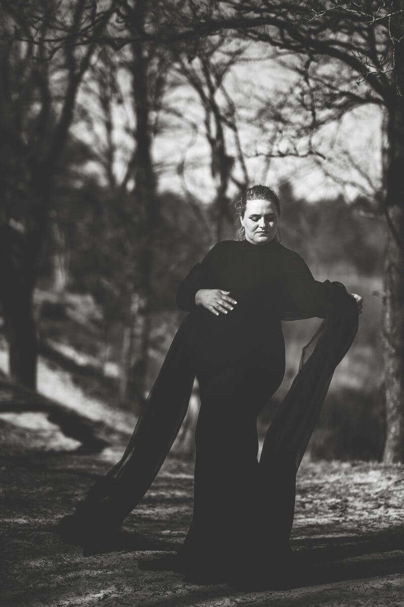Zwangere vrouw in lange zwarte jurk op bospad tijdens zwangerschapsfotoshoot, met kale bomen en onscherpe achtergrond. Elegante, dramatische zwart-wit foto van zwangerschapsshoot in de natuur.