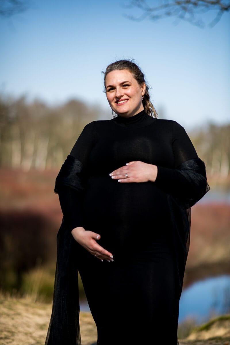 Vrouw poseert buiten tijdens zwangerschapsfotoshoot in lange zwarte jurk, hand op buik, lastig. Natuurlijk decor met bomen, gras en blauwe lucht op de achtergrond.