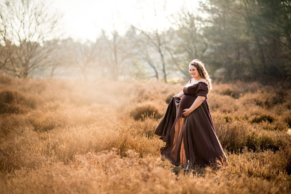 Zwangerschapsfotoshoot in krachtig veld met hoge, gouden grasprieten. Zwangere vrouw in bruine jurk houdt buik vast, omringd door kale bomen en constante lucht voor serene sfeer.