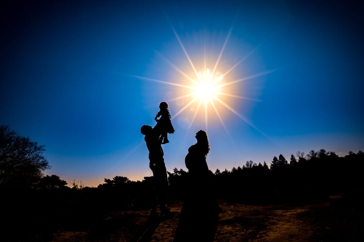 Gezin tijdens zwangerschapsfotoshoot, moeder tilt soort op bij krachtig weer, silhouetten duidelijk zichtbaar tegen blauwe lucht, bomen en struiken op de achtergrond.