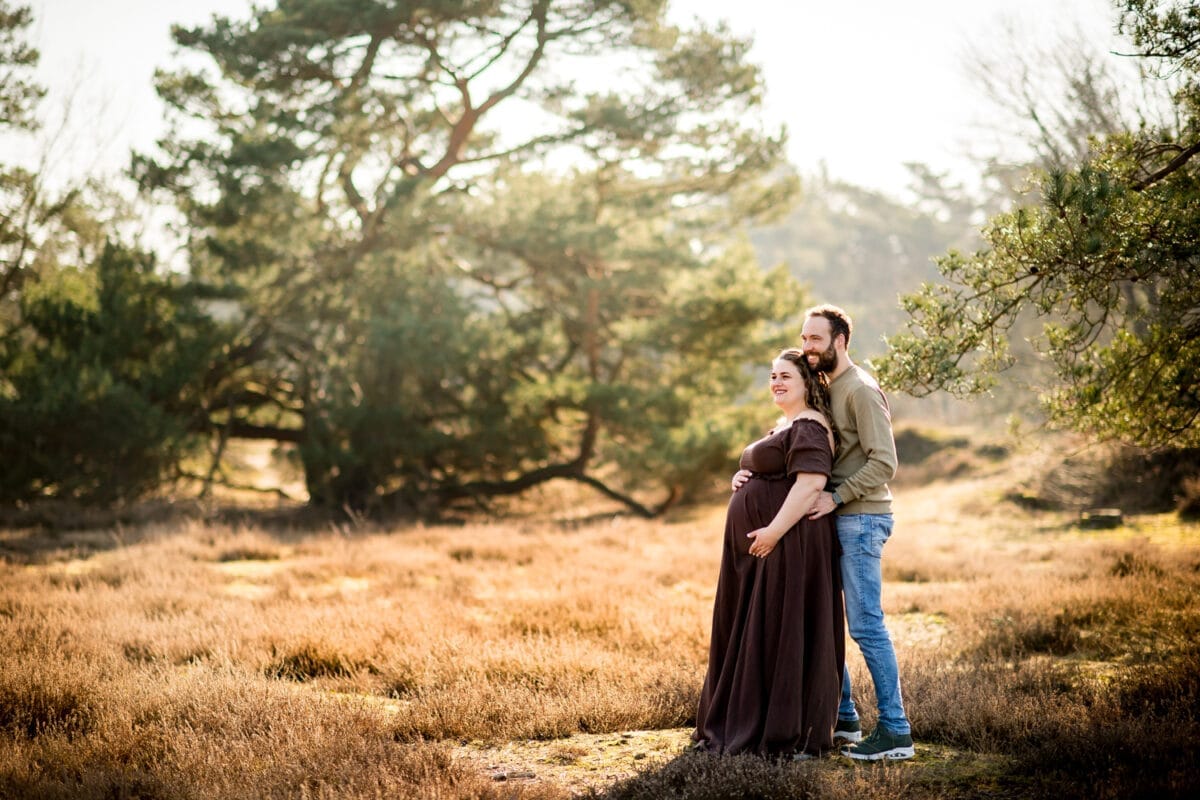Stel omhelst zwangere vrouw in krachtig veld, romantische zwangerschapsfotografie met groene bomen, bruine jurk en warme zonnestralen. Buik wordt liefdevol.