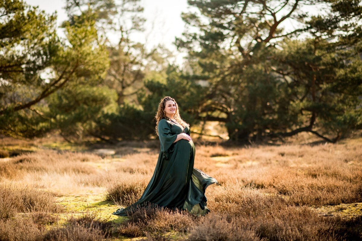 Zwangerschapsfotoshoot in zonovergoten veld met zwangere vrouw in donkergroene jurk, omgeven door natuur, grote bomen en droge gras. Natuurlijke, serene zwangerschapsfotografie buiten.