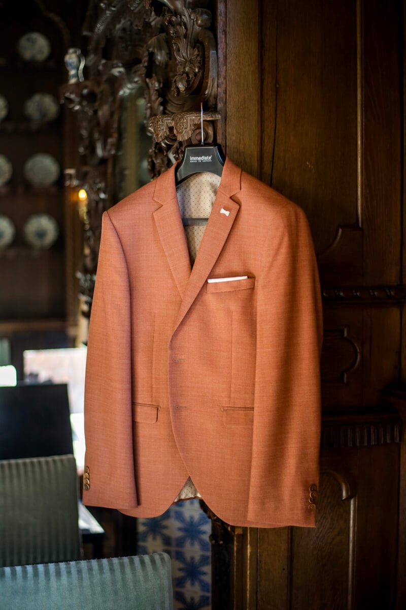 Stijlvolle oranje colbert met wit pochet op hanger tegen donkere houten wand bij Nienoord. Vintage sfeer, kleine houtsnijwerk en decoratieve borden, ideale trouwlocatie decoratie.