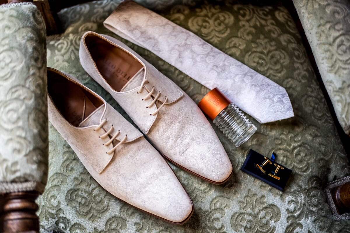 Luxe beige herenschoenen, witte stropdas met patroon, parfumflesje en gouden manchetknopen op groene stoel – elegante bruiloftaccessoires voor heren.