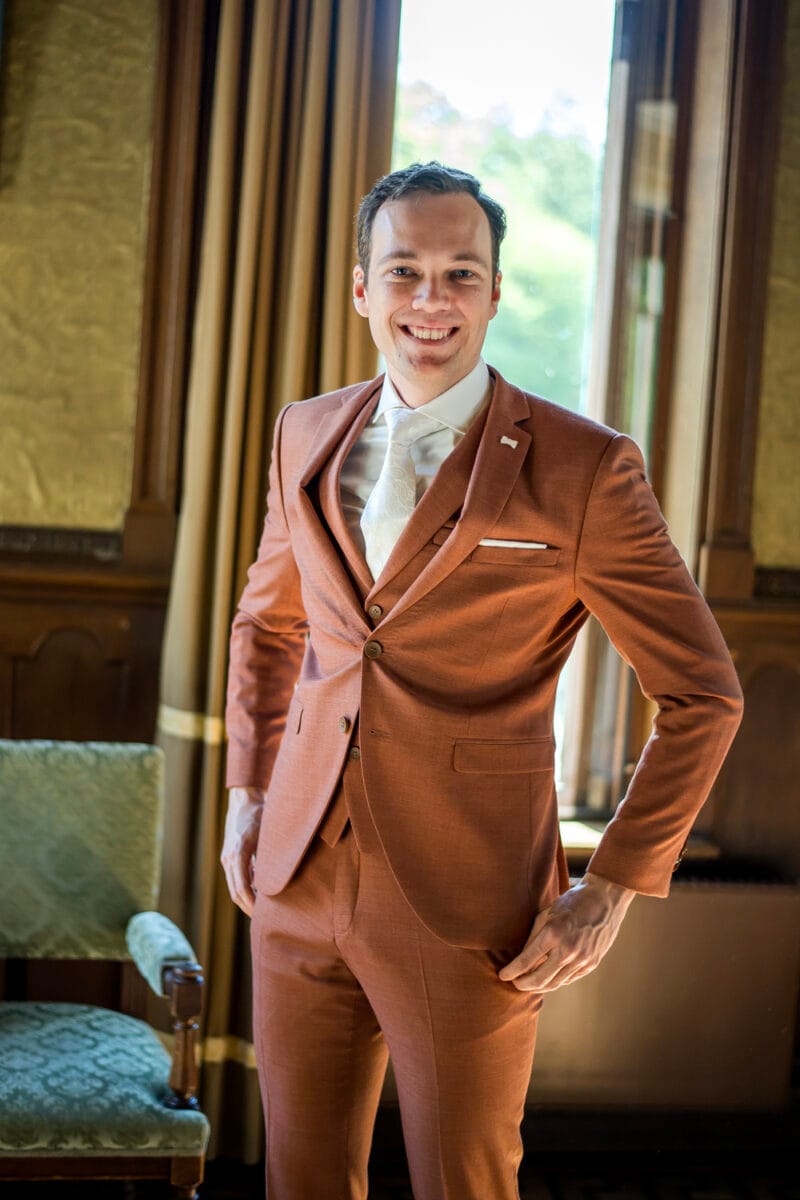 Man in roestkleurig pak poseert bij groene stoel voor grote ramen met gouden gordijnen tijdens bruiloft Nienoord in lichte binnenruimte. Foto bruiloft Nienoord interieur, luxe sfeer.