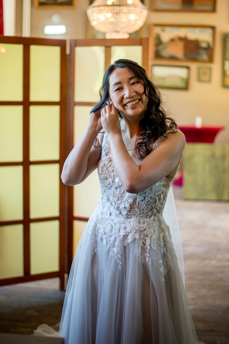 Bruid in kanten mouwloze trouwjurk met gekruld donker haar past oorbellen aan op de trouwlocatie, gele schermen en ingelijste foto's op de achtergrond. Het moment van het voorbereiden van de bruidssieraden.