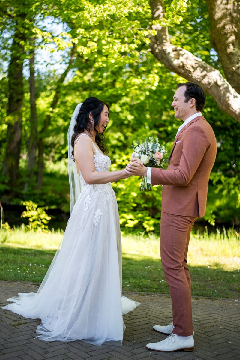 Bruid in witte jurk en bruid in roestkleurig pak lachend samen hand in hand tijdens buitenbruiloft op Nienoord, omringd door groene bomen op een zonnige dag.