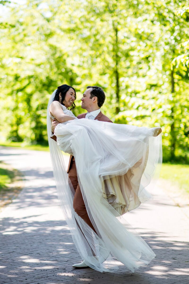 Bruiloft op Nienoord: bruid tilt bruid in witte trouwjurk en sluier op sterke bospad, omgeven door groene bomen. Romantische huwelijksfoto's in de natuur.