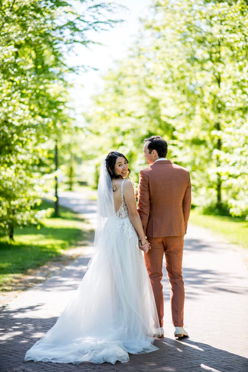 Bruid in witte jurk en sluier naast bruid in roestkleurig pak op zonnige bruiloft bij Landgoed Nienoord, omringd door groene bomen, lachend kijkend naar de camera.