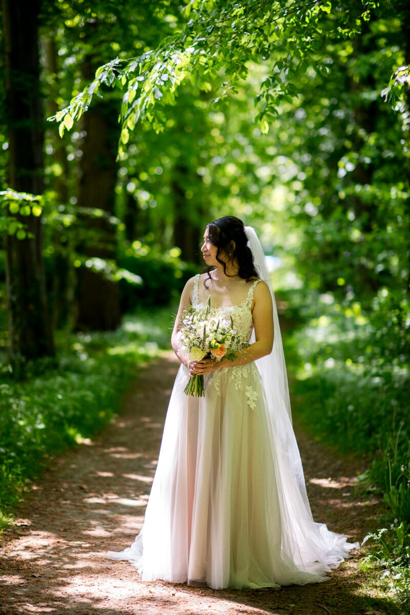 Bruiloft Nienoord bruid in witte kanten trouwjurk met lange sluier en bloemenboeket, staand op zonovergoten pad omgeven door bomen en natuur, romantische trouwfoto buiten.