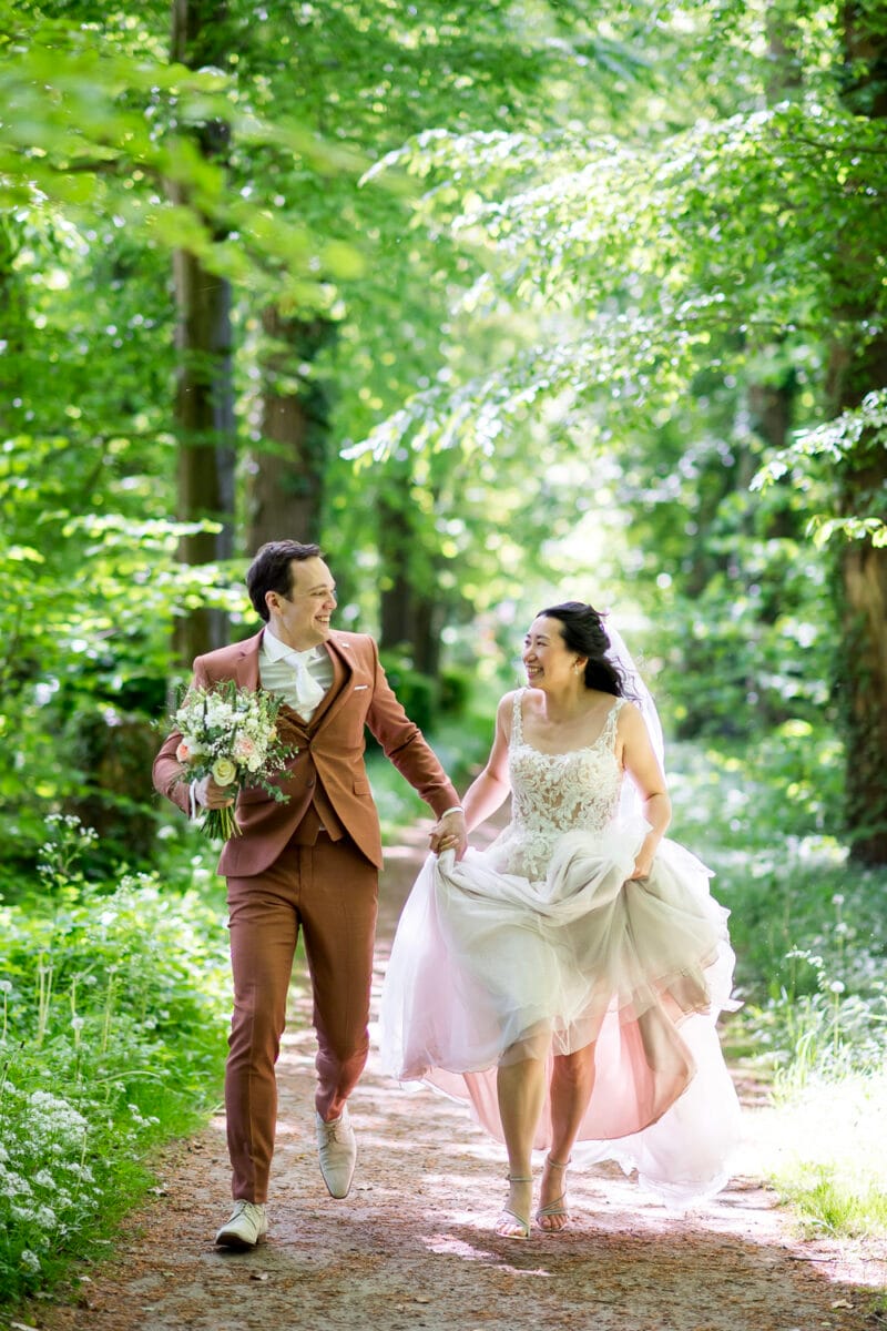 Bruiloft Nienoord: bruid in witte jurk en bruid in roestkleurig pak rennen hand in hand over zonovergoten bospad, met boeket, groene bladeren en gouden zonlicht.