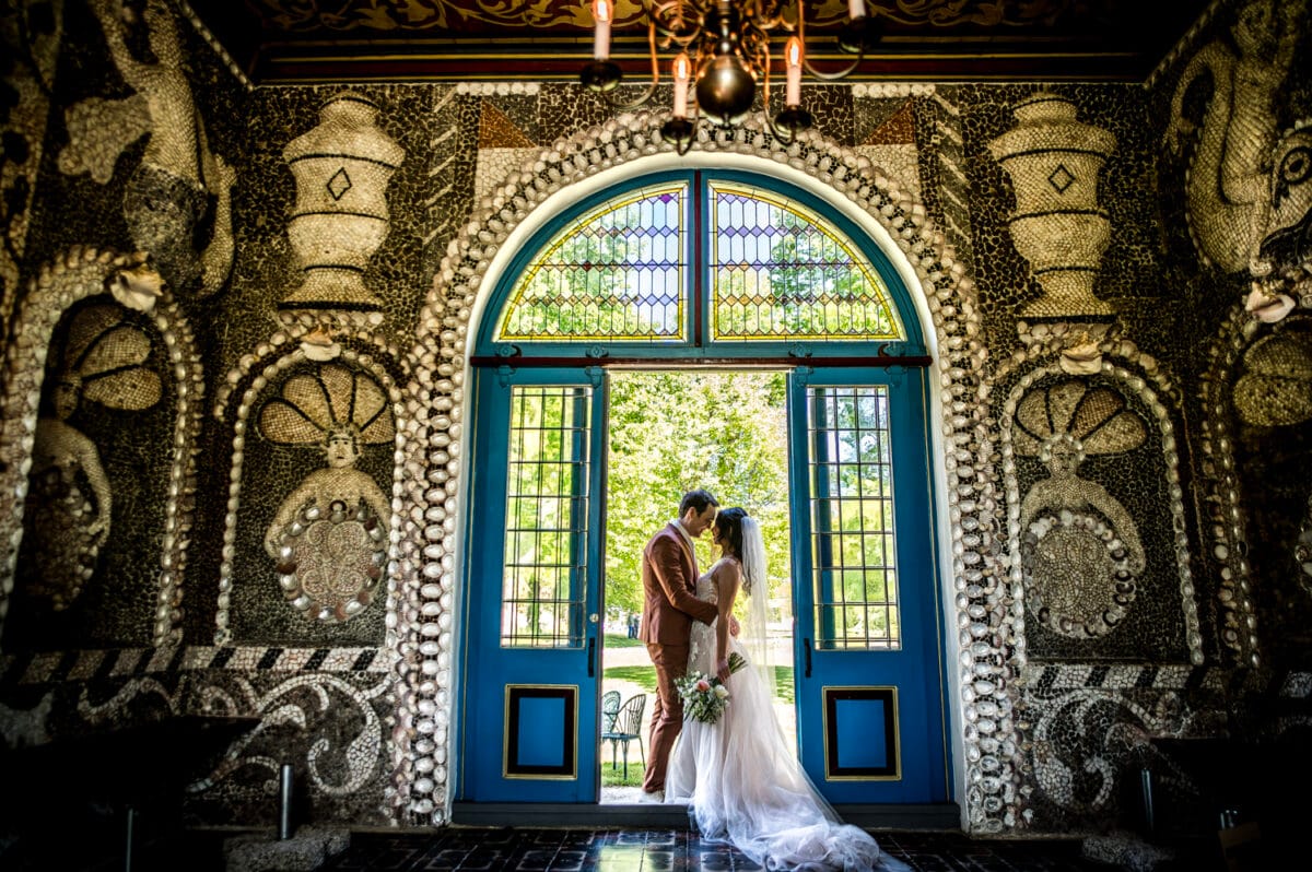 Bruid en bruidegom omhelzen elkaar voor grote blauwe boogdeuren met glas-in-lood bij Bruiloft Nienoord, verborgen door schelpendecoratie en zonlicht op de witte trouwjurk en boeket.