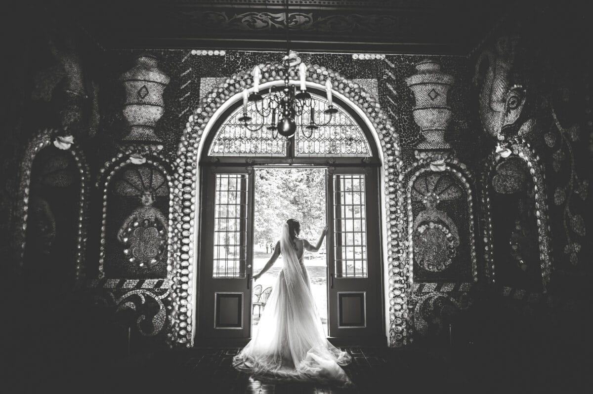 Bruid in elegante trouwjurk op landgoed Nienoord, zonlicht door open deuren, sierlijke kamer met schelpen- en mozaïekdecoratie aan de muur, kroonluchter erboven. Zwart-wit trouwfoto.
