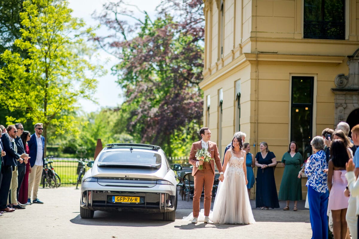 Bruiloft bij Landgoed Nienoord: bruid in witte jurk en bruid in roestkleurige pak staan hand in hand buiten voor gele villa, met gasten, Nederlandse auto, fietsen en groene bomen op de achtergrond.