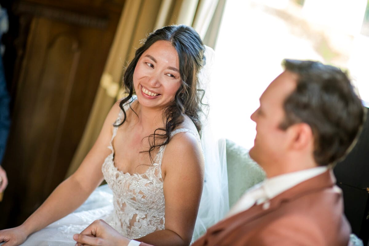 Bruiloft Nienoord: bruid in witte kanten jurk en sluier lacht naar bruid in bruin pak. Intiem, onzichtbaar moment bij raam op hun trouwdag.