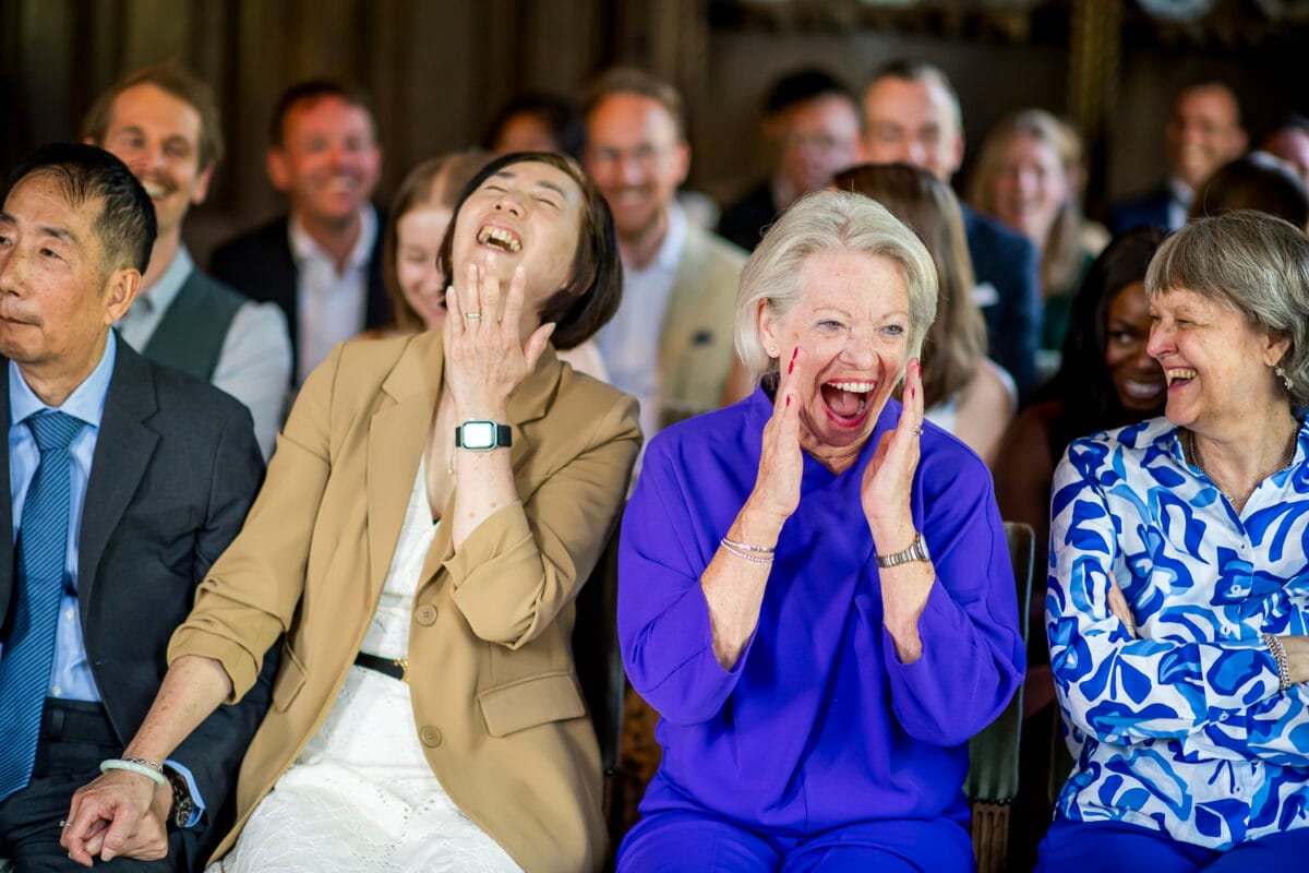 Bruiloftsfeest op Nienoord met lachende gasten. Twee vrouwen vooraan, één in beige en één in felblauw, lachend uitbundig. Sfeervolle trouwfoto vol vrolijkheid en plezier.