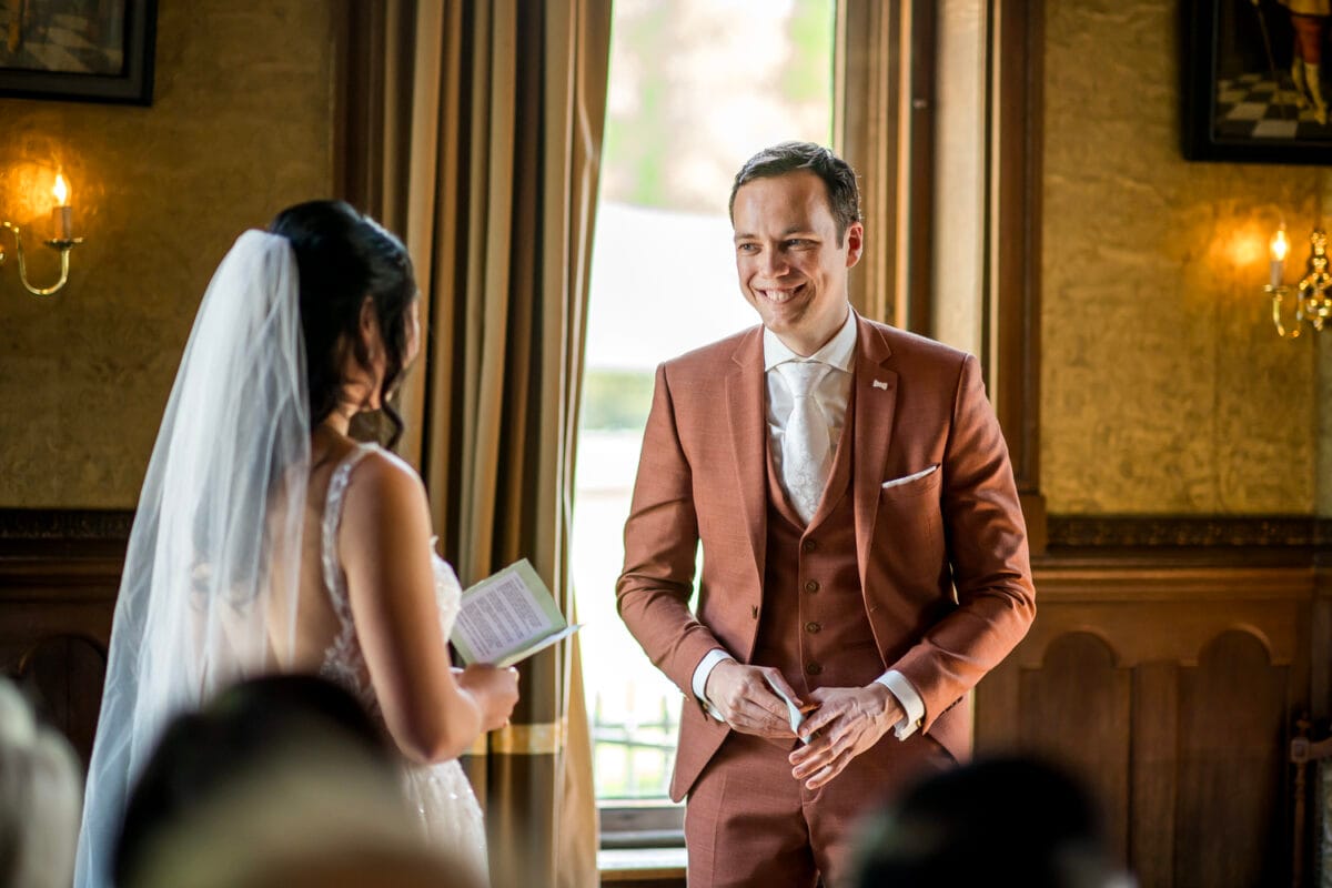 Bruiloft bij Nienoord: bruid in roestkleurige pak en bruid in witte trouwjurk met sluier bij raam, elegante kamer met houten lambrisering en gouden gordijnen.