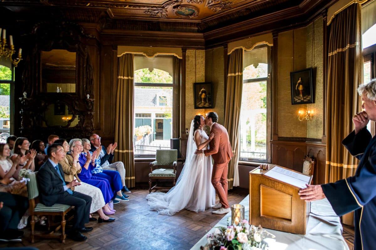 Bruidspaar kust aan altaar in elegante, houtbeklede zaal op Landgoed Nienoord tijdens bruiloft; gasten klappen en lachen terwijl trouwambtenaar ceremonie leidt tot zonlicht.