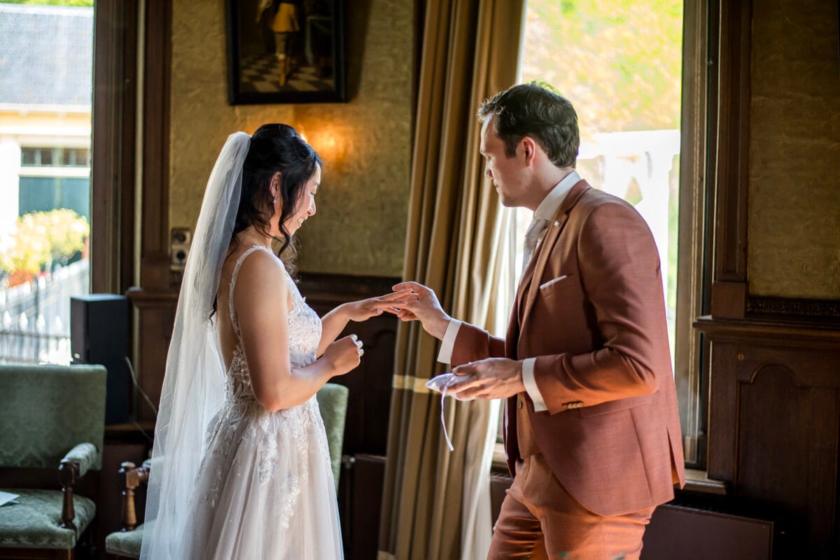 Bruiloft Nienoord: bruid in bruin pak schuift trouwring om vinger van bruid bij vaste raam, omgeven door groene stoelen en goudkleurige gordijnen in sfeervol verlichte trouwzaal.