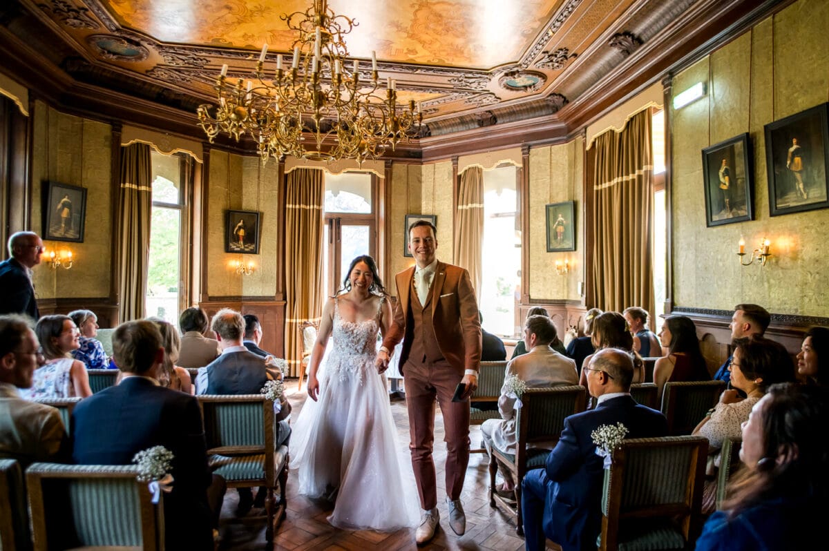 Bruiloft ceremonie Nienoord: bruidspaar loopt hand in hand het gangpad af in luxe zaal met gouden plafonds, kroonluchters en grote ramen, omringd door ronde gasten.