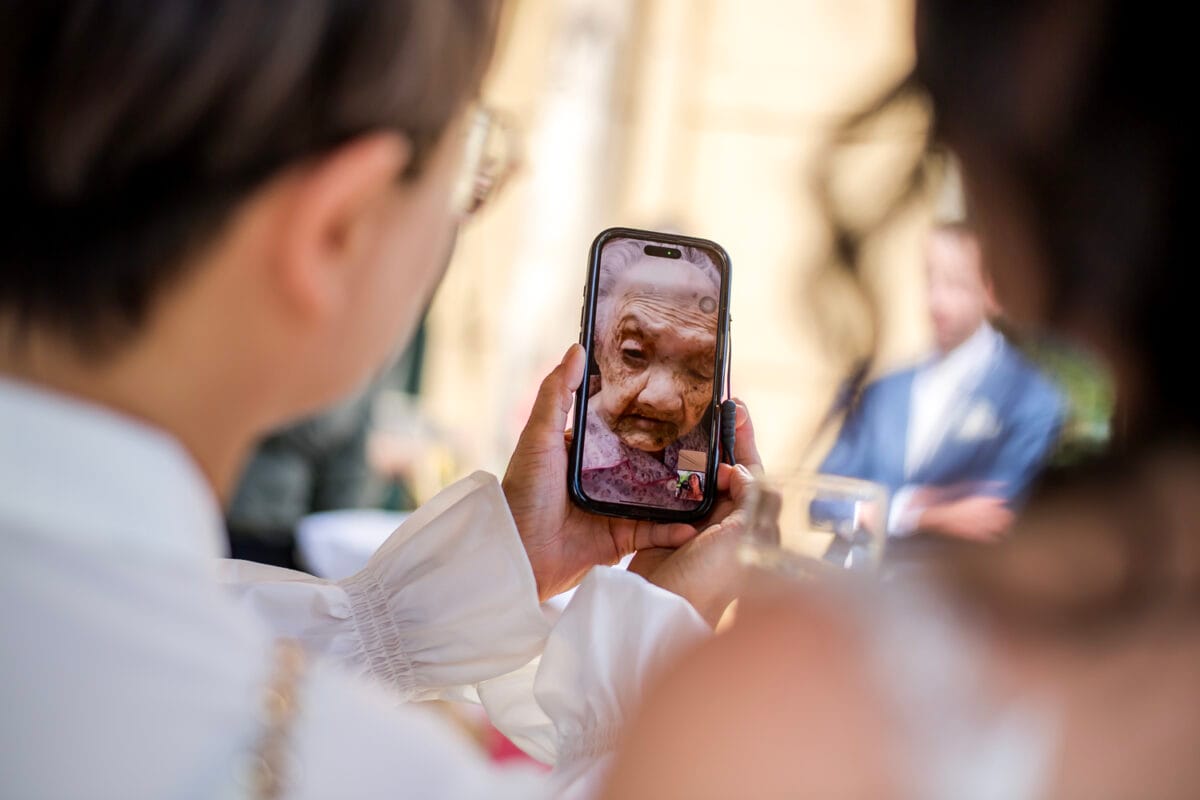 Smartphone met videogesprek op bruiloft Nienoord, ouderen op afstand, gasten in feestkleding buiten tijdens huwelijksceremonie Chi Jan en Jesse.