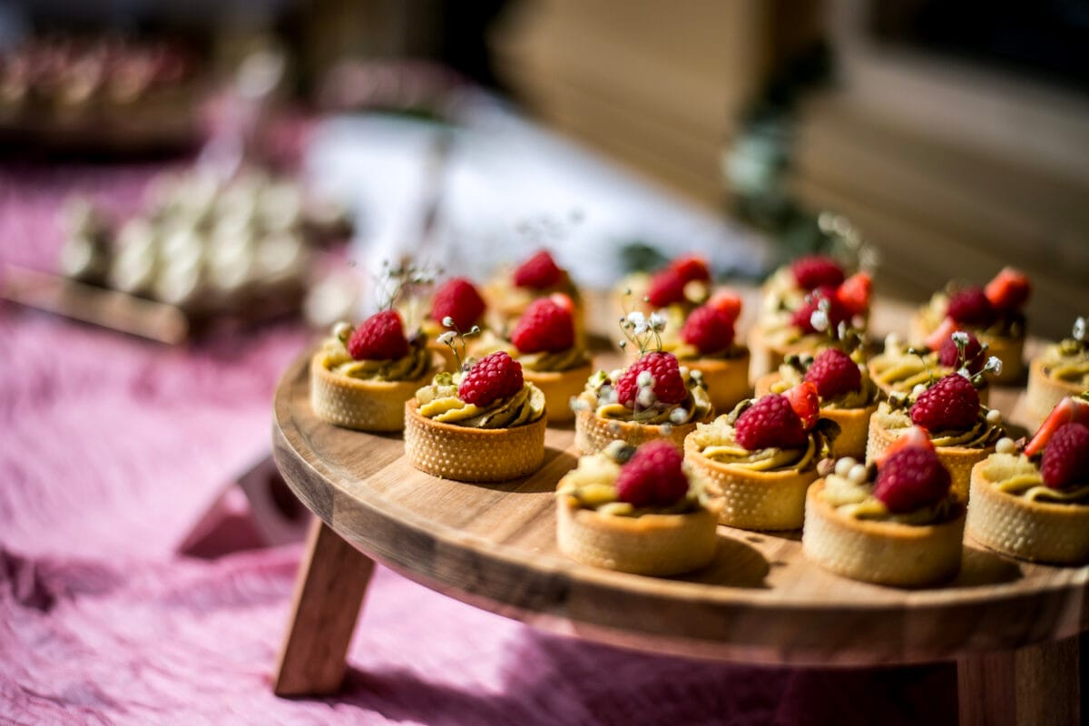 Houten dienblad met mini fruittaartjes met verse frambozen en witte bloemen, stijlvolle stijlvolle op roze tafellaken tijdens bruiloft in Nienoord, dessert catering inspiratie.