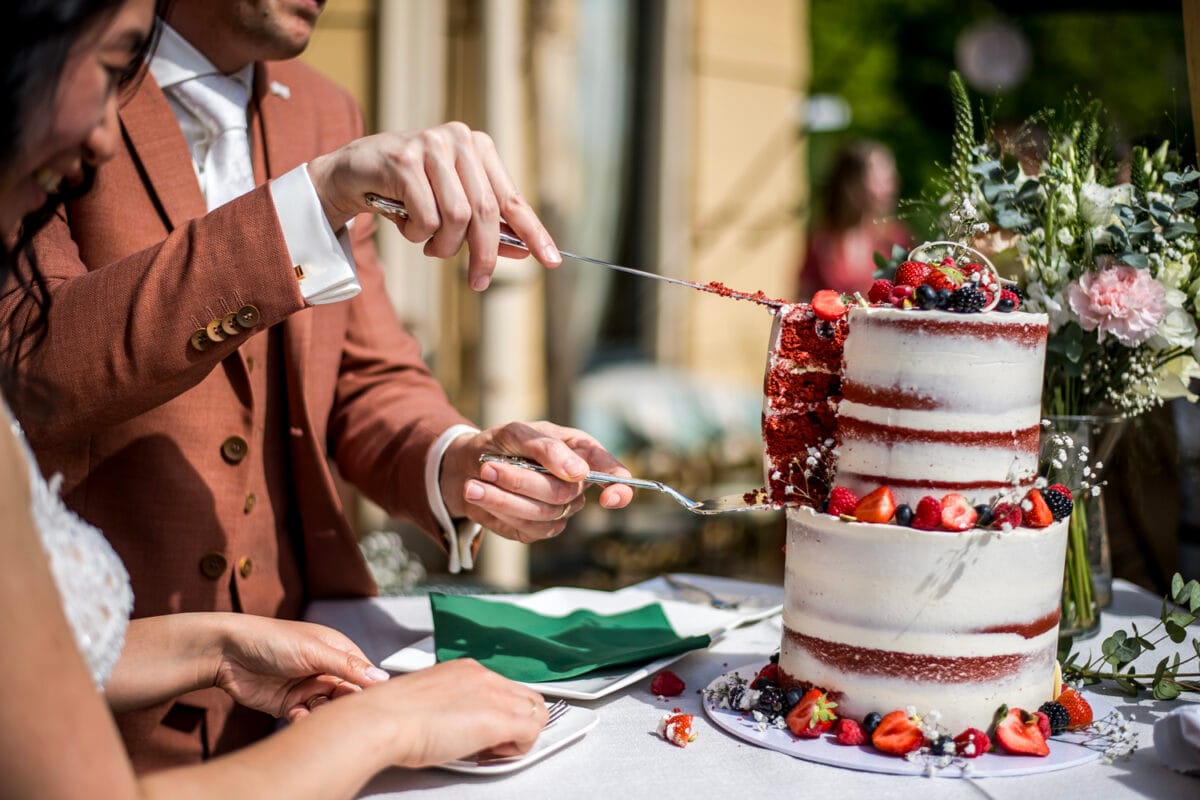Bruiloft Nienoord bruid en bruidegom snijden semi-naked tweelaagse bruidstaart met bessen en bloemen buiten, omringd door groene en bloemrijke decoratie in zonlicht.