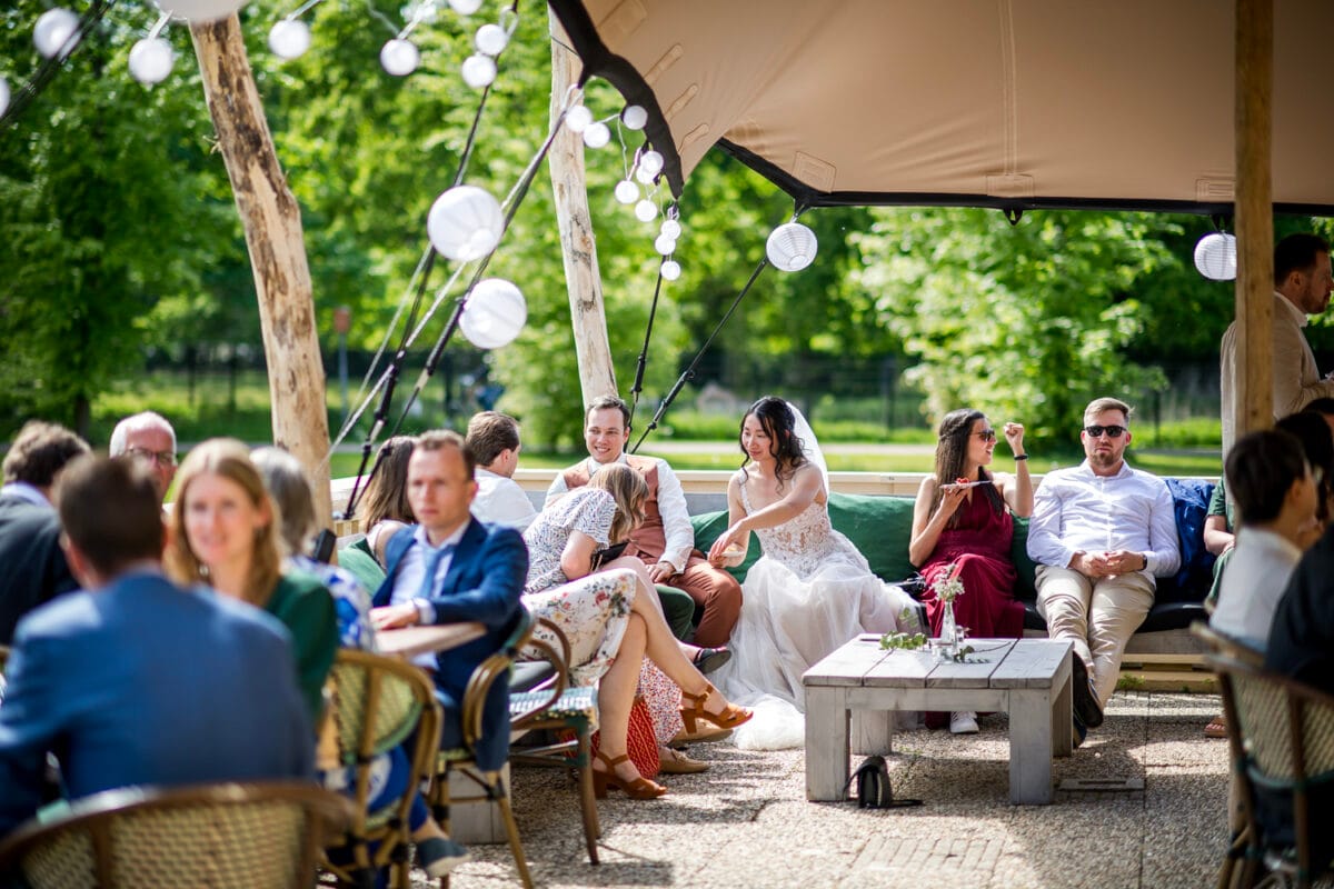 Bruiloft in de buitenlucht op Nienoord met bruid in witte jurk, gasten in formele kleding, houten tafels, witte lantaarns onder een afdak, omringd door groene bomen. Bruiloftsreceptie.