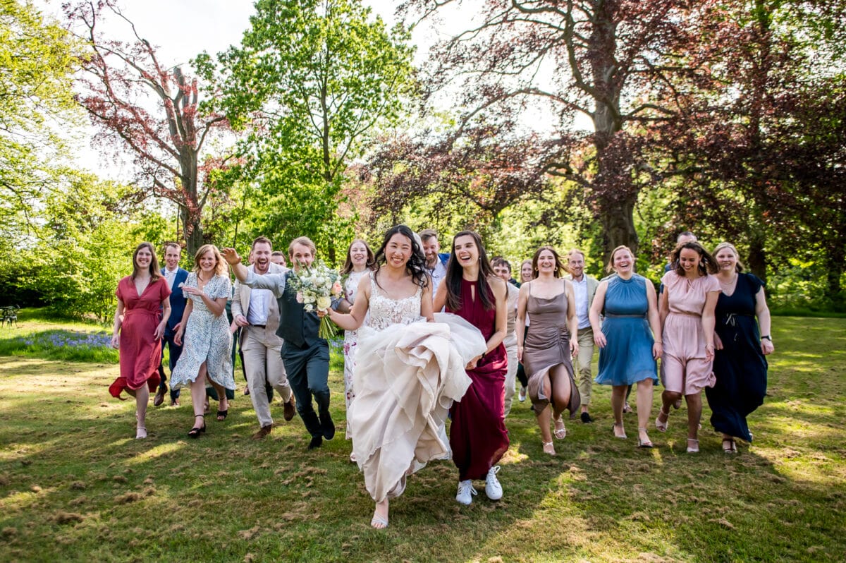 Bruid rent met boeket over zonnig gazon op trouwlocatie Nienoord, omgeven door bruidsgezelschap, hoge bomen en weelderig groen in een feestelijke buitensetting.