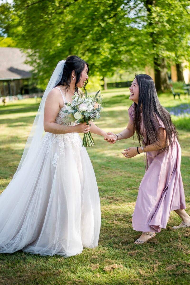 Bruid in witte jurk en vrouw in mauve jurk lachen samen buiten bij Bruiloft Nienoord, zonlicht door bladeren, vrolijke bruiloft sfeer. Buitenbruiloft fotografie locatie Nederland.