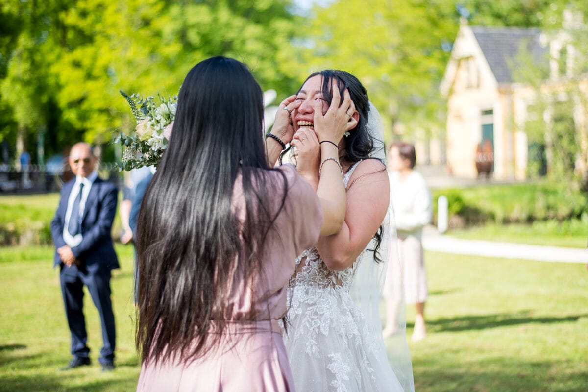 Bruiloft bij Nienoord: bruid in witte kanten jurk met boeket krijgt liefdevol tranen afgedroogd door vrouw in roze jurk, omringd door zonlicht, bomen en gemengde geklede gasten.