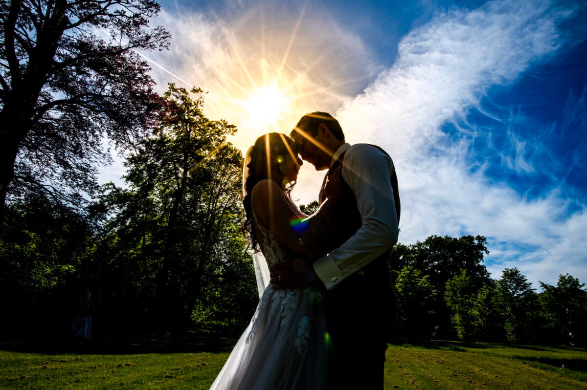 Trouwfoto buiten bij Nienoord: bruidspaar omhelst elkaar onder zonnestralen, silhouet tegen blauwe lucht met groene bomen en lichte wolken, romantische sfeer op zonnige dag.