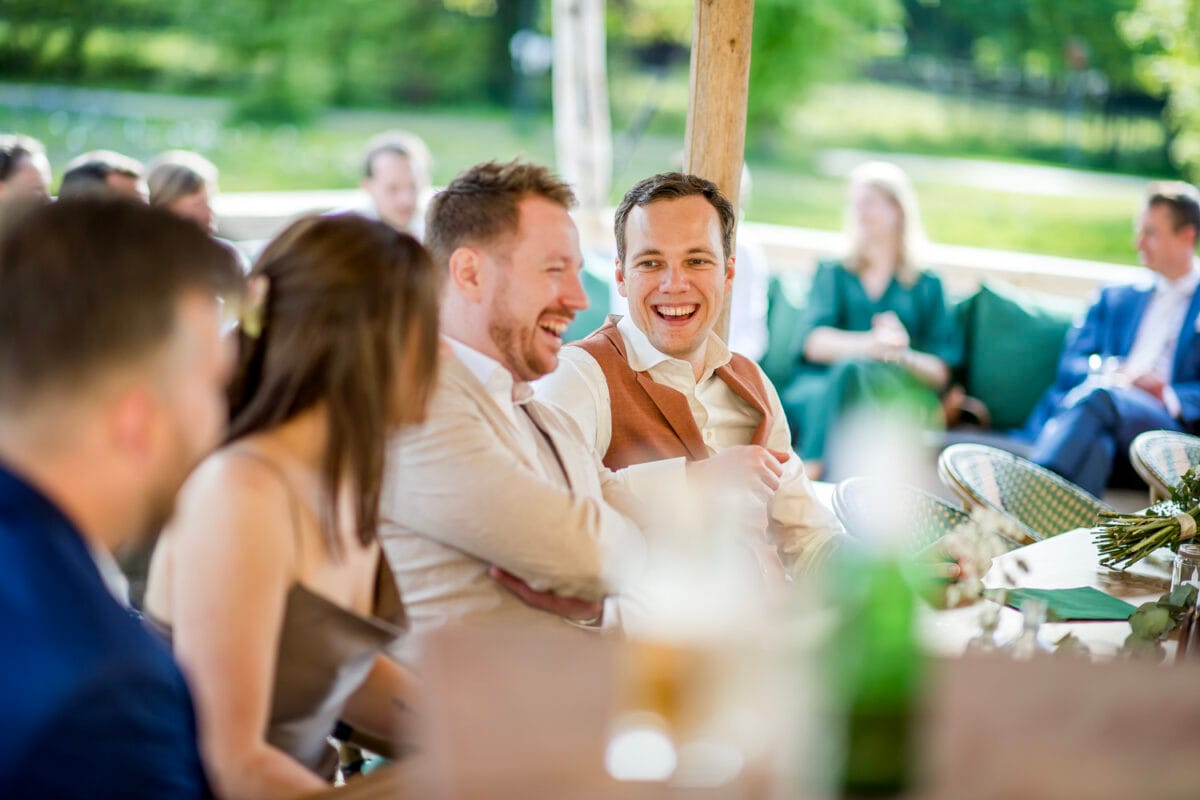 Gezellige bruiloft op Nienoord: groep mensen aan buitentafel, lachend en pratend in zonlicht onder groene bomen, feestelijke sfeer tijdens huwelijksreceptie.