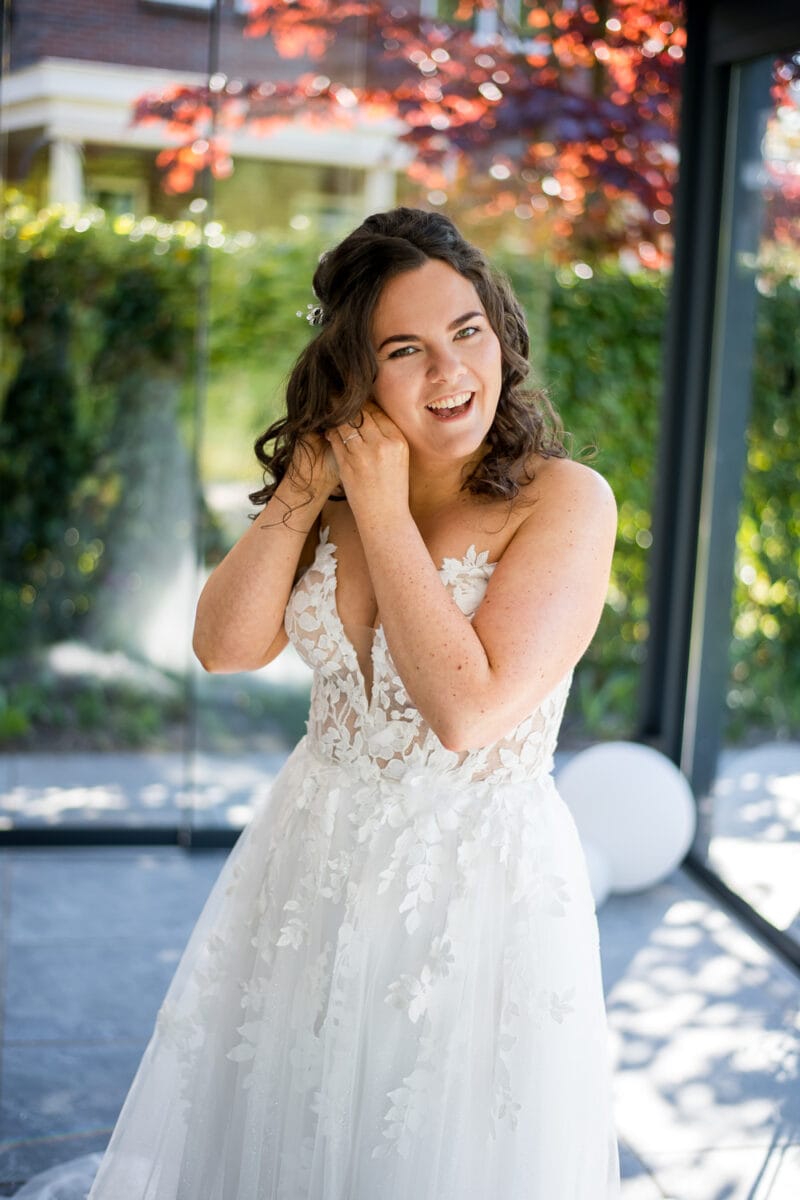 Bruid in witte kanten trouwjurk doet oorbel in bij Garnwerd aan Zee, met zonlicht en uitzicht op groen en rode bladeren door grote ramen. Bruiloft binnen, romantische sfeer.