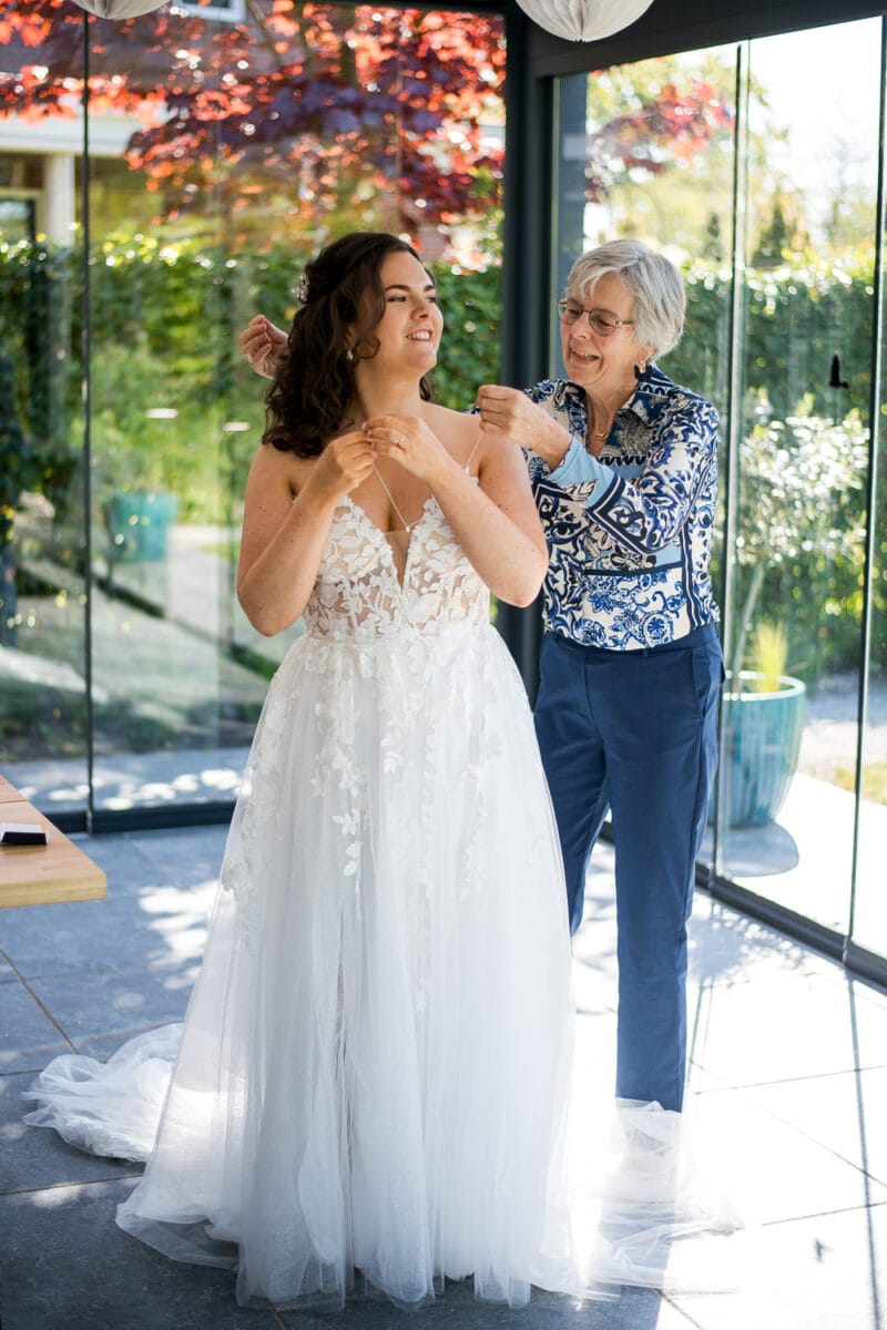 Bruiloft bij Garnwerd aan Zee: bruid in witte kanten trouwjurk krijgt hulp met jurkbandjes door oudere vrouw, binnen in zonnige ruimte met grote ramen en uitzicht op groen.