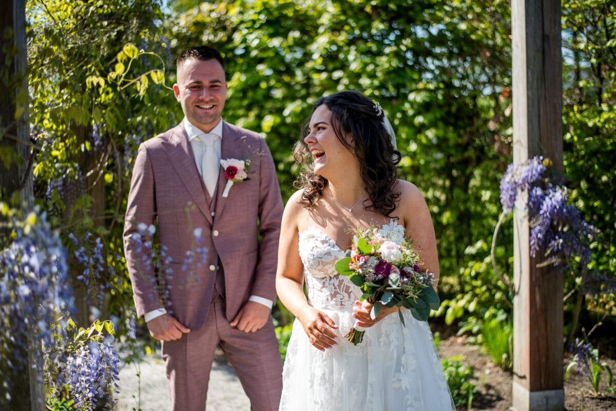 Bruid in witte kanten jurk en bruid in lichtroze pak lachend op buitenbruiloft bij Garnwerd aan Zee, omringd door groene natuur en paarse bloemen onder zonnige lucht.
