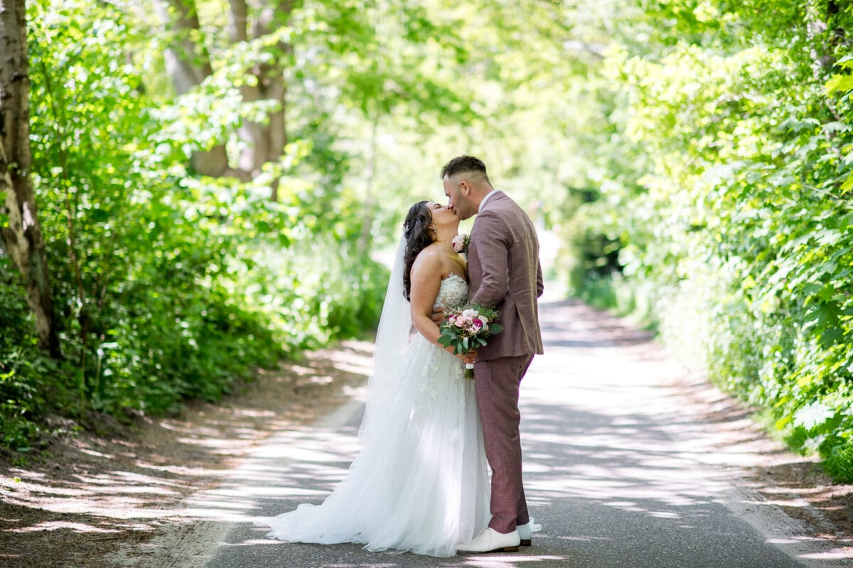 Romantische bruiloft nabij Garnwerd aan Zee: bruid en bruidegom kussen op zonovergoten weg, omringd door groene bomen. Bruid in witte jurk, bruid in lichtbruin pak.