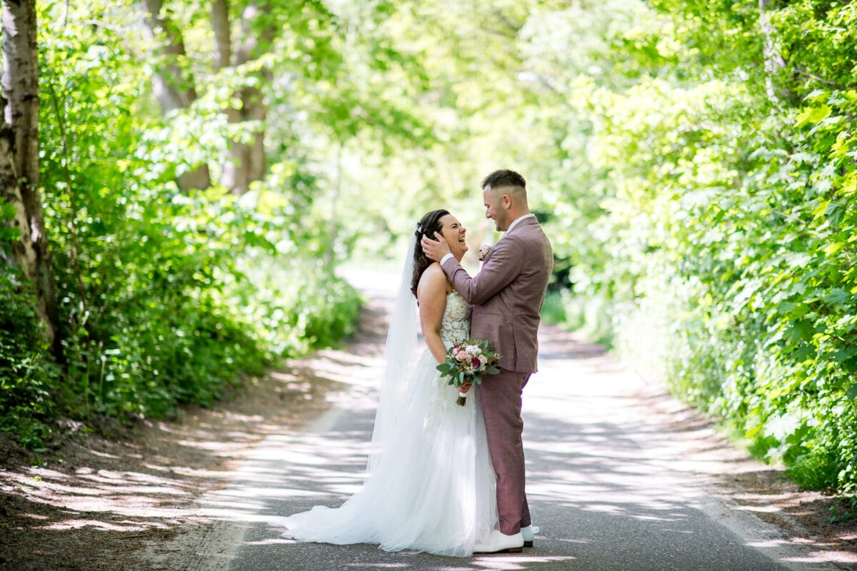 Bruidspaar op zonovergoten bospad tijdens bruiloft bij Garnwerd aan Zee, bruid in witte jurk, bruid in licht mauve pak, omringd door groen en romantische sfeer.