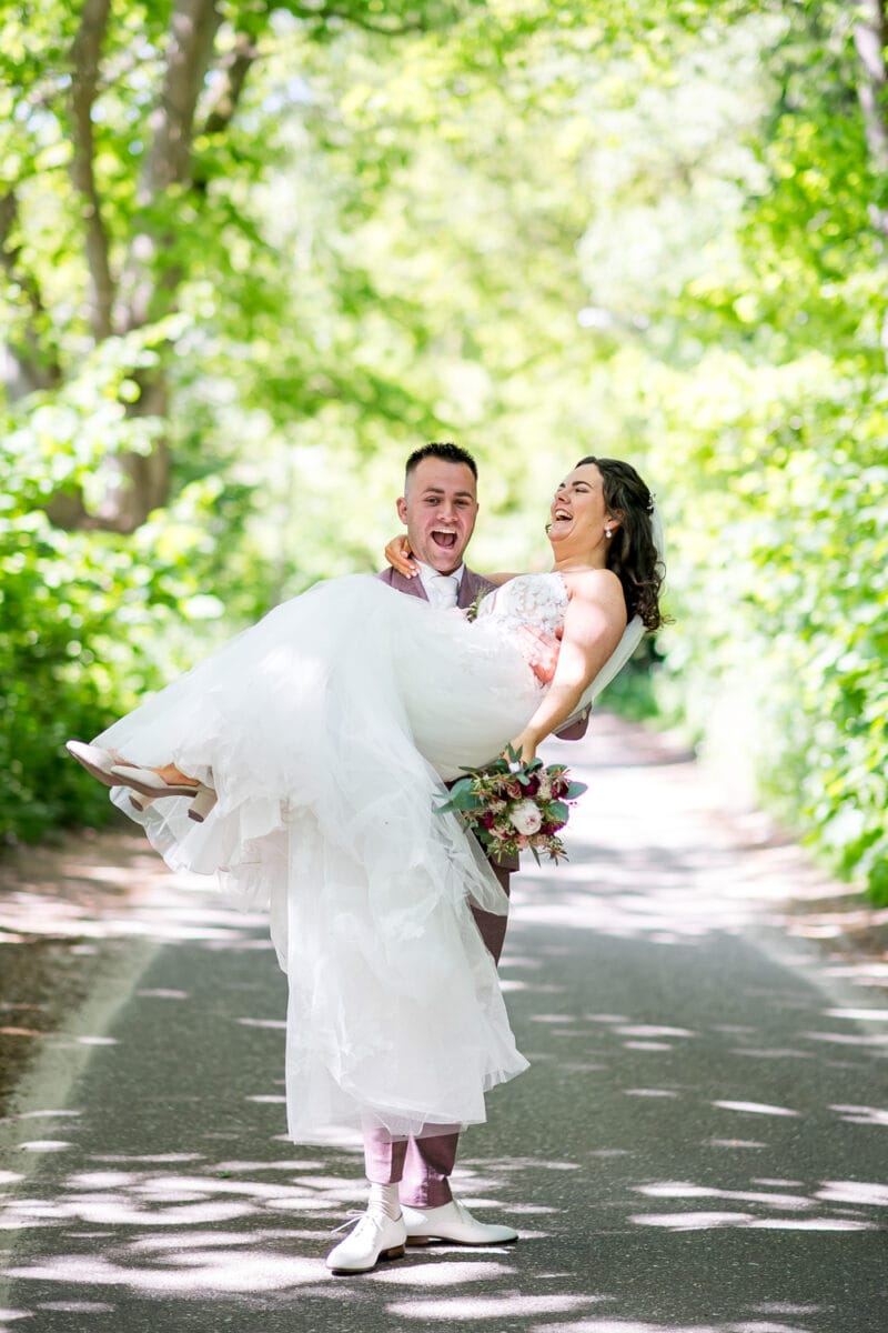 Bruiloft bij Garnwerd aan Zee: bruid tilt bruid in witte jurk, kleurrijk boeket, zonlicht en groene bladeren, vrolijk en zorgeloos stel op bosrijk trouwpad.