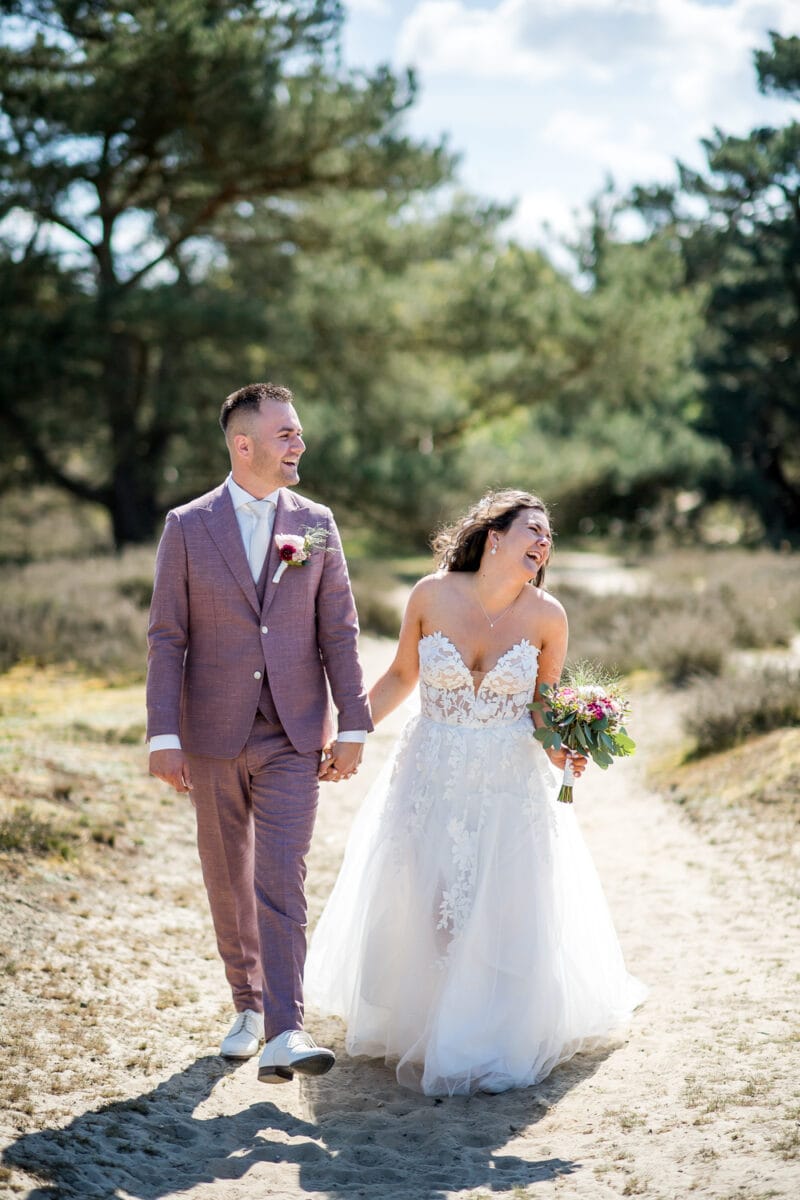 Bruid in witte kanten strapless trouwjurk en bruid in mauve pak wandelen hand in hand op zonnige bruiloft bij Garnwerd aan Zee, tussen groen en zandpad, lachend en ontspannen.