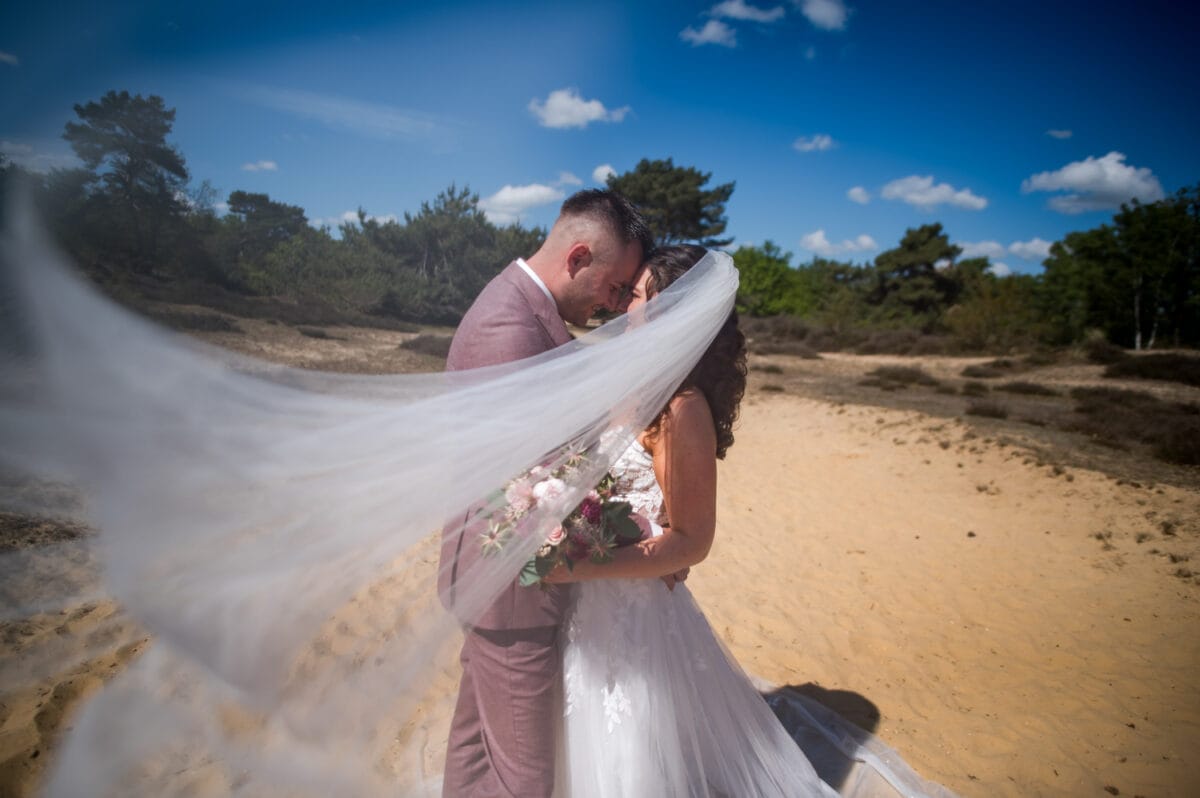 Bruid en bruidegom op het strand van Garnwerd aan Zee met blauwe lucht, wapperende sluier en boeket bloemen; romantische bruiloftsfoto's in natuurlijke omgeving.