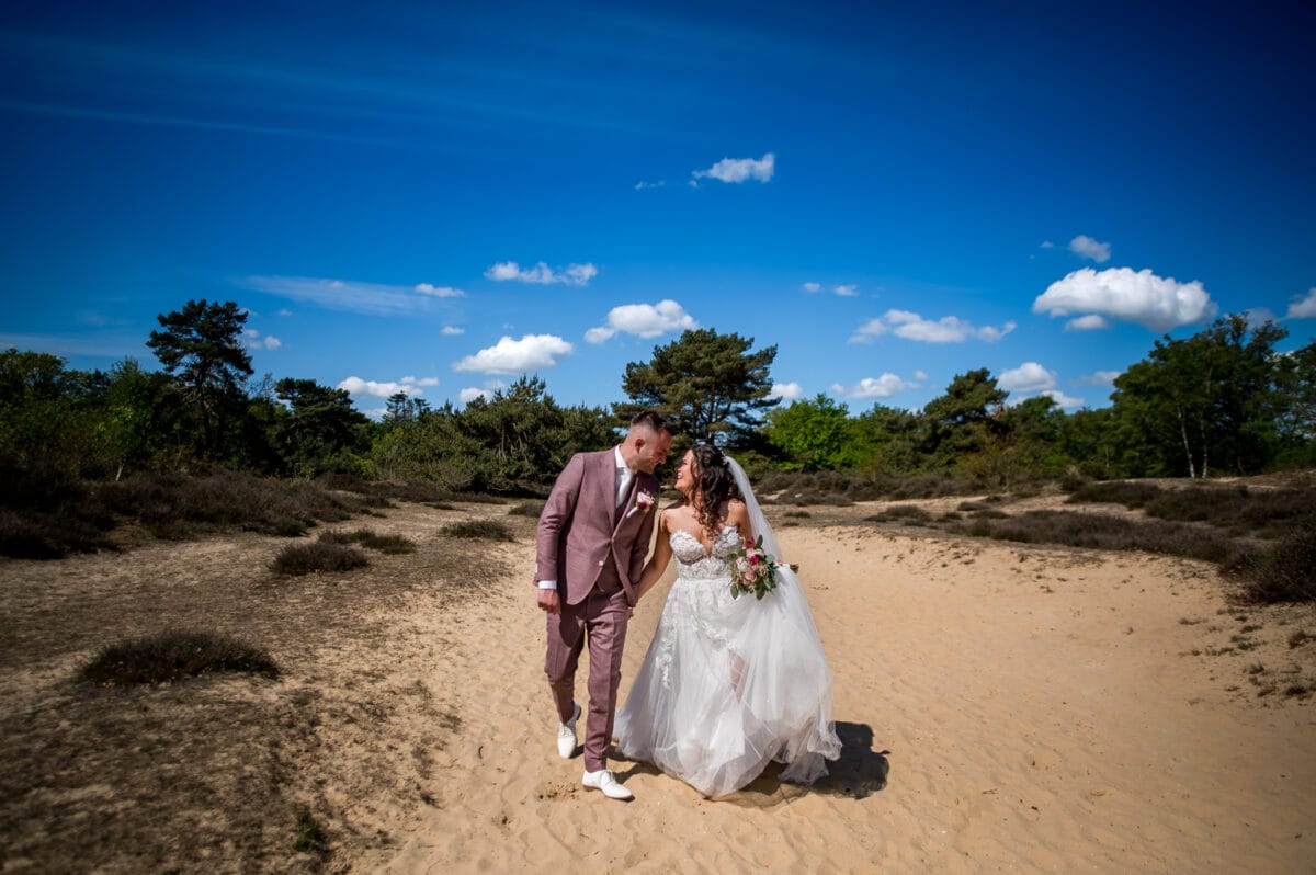 Bruid en bruidegom op zandpad bij Garnwerd aan Zee, omgeven door groene bomen en struiken, genieten van hun romantische bruiloft onder blauwe lucht met wolken.
