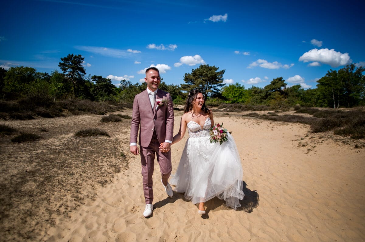 Bruiloftspaar loopt hand in hand op zandgrond onder blauwe lucht bij Garnwerd aan Zee, bruid in witte strapless trouwjurk en bruid in lichtroze pak, omringd door struiken en bomen.