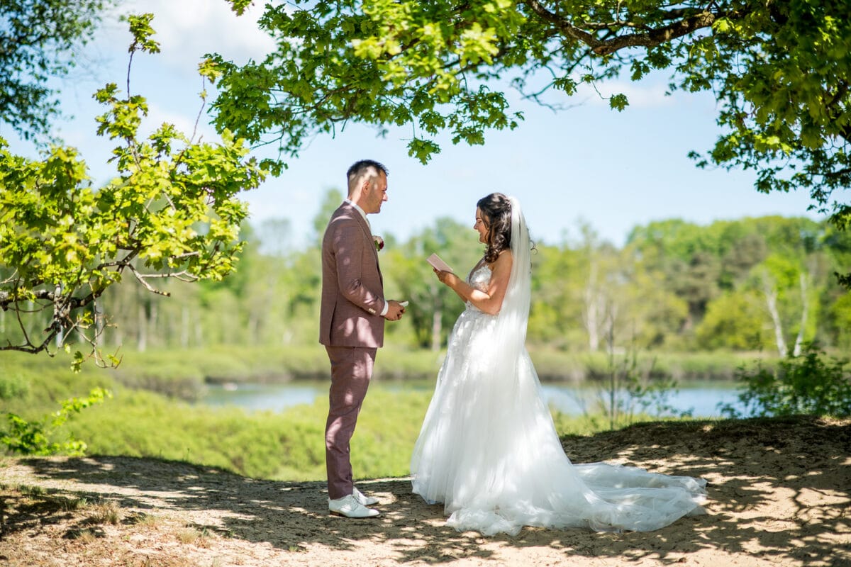 Bruid leest geloften voor aan bruid bij Garnwerd aan Zee bruiloft, buiten in de zon met vijver, groene bomen en blauwe lucht op de achtergrond. Romantische trouwfoto in de natuur.